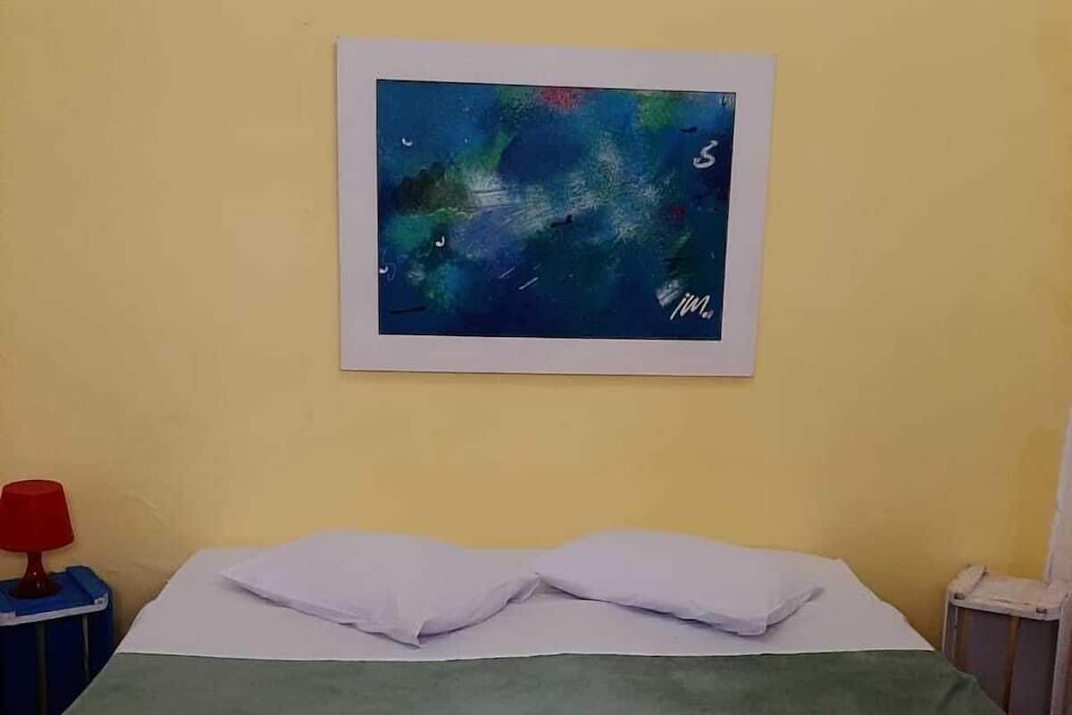Um quarto com uma cama, um quadro na parede, um abajur vermelho e um banquinho azul.