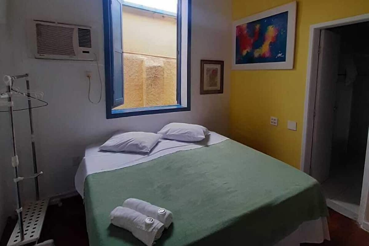 Quarto com cama, janela, ar condicionado fixado na parede, porta e um quadro colorido.