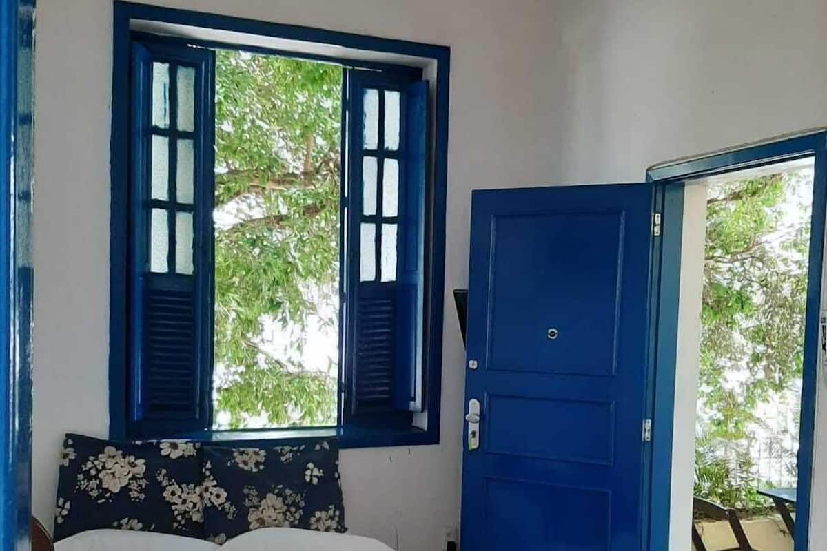 Quarto com cama, janela com persianas, porta azul e vista para o verde lá fora.