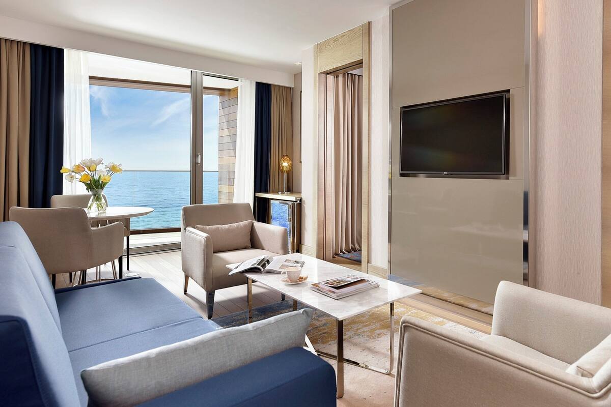 A hotel room with a sofa, armchairs, a coffee table, a flat-screen TV, and a balcony with a view of the sea.