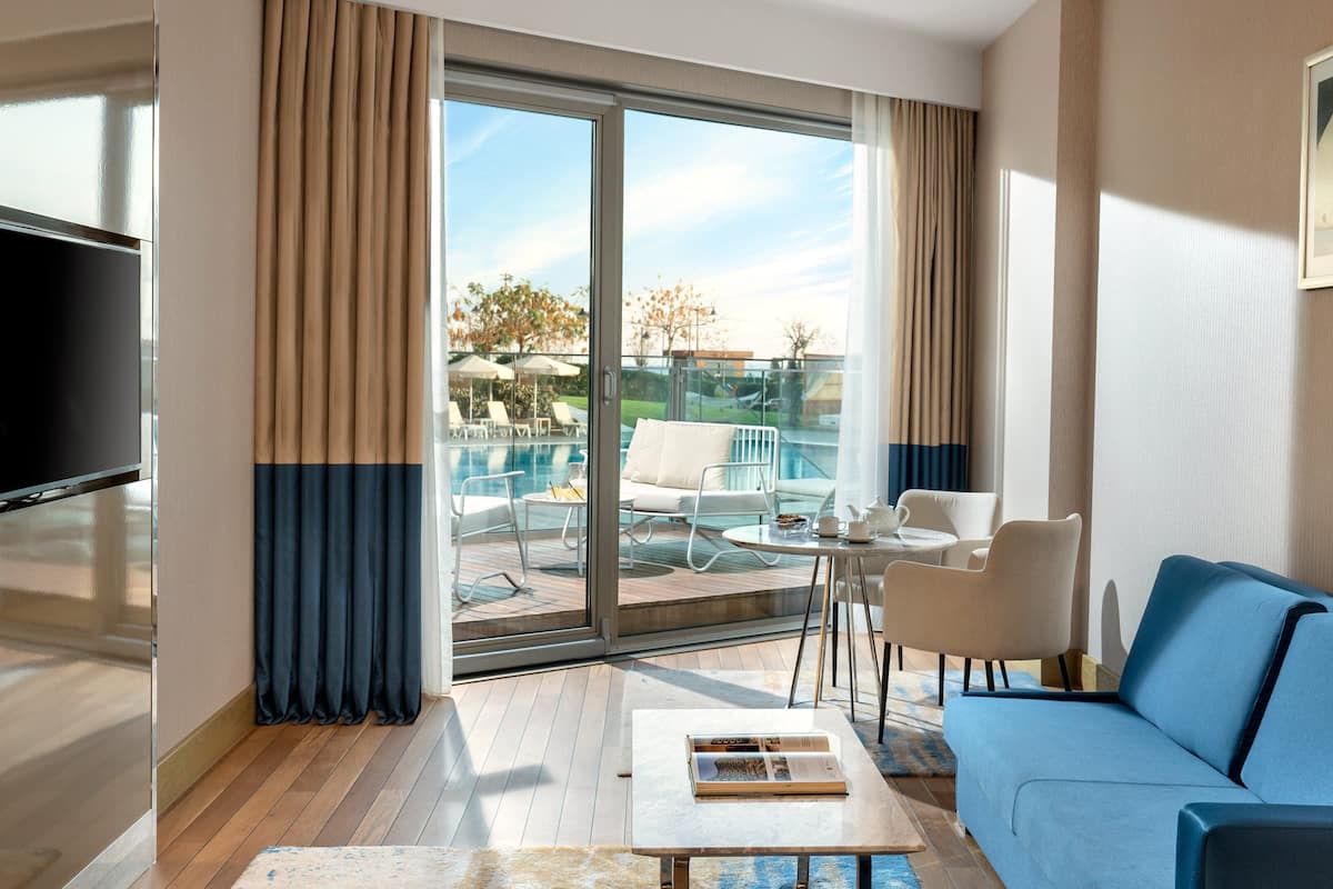 A modern hotel room with a large sliding glass door leading to a pool area, a flat-screen TV, a blue sofa, and a small table with a book.