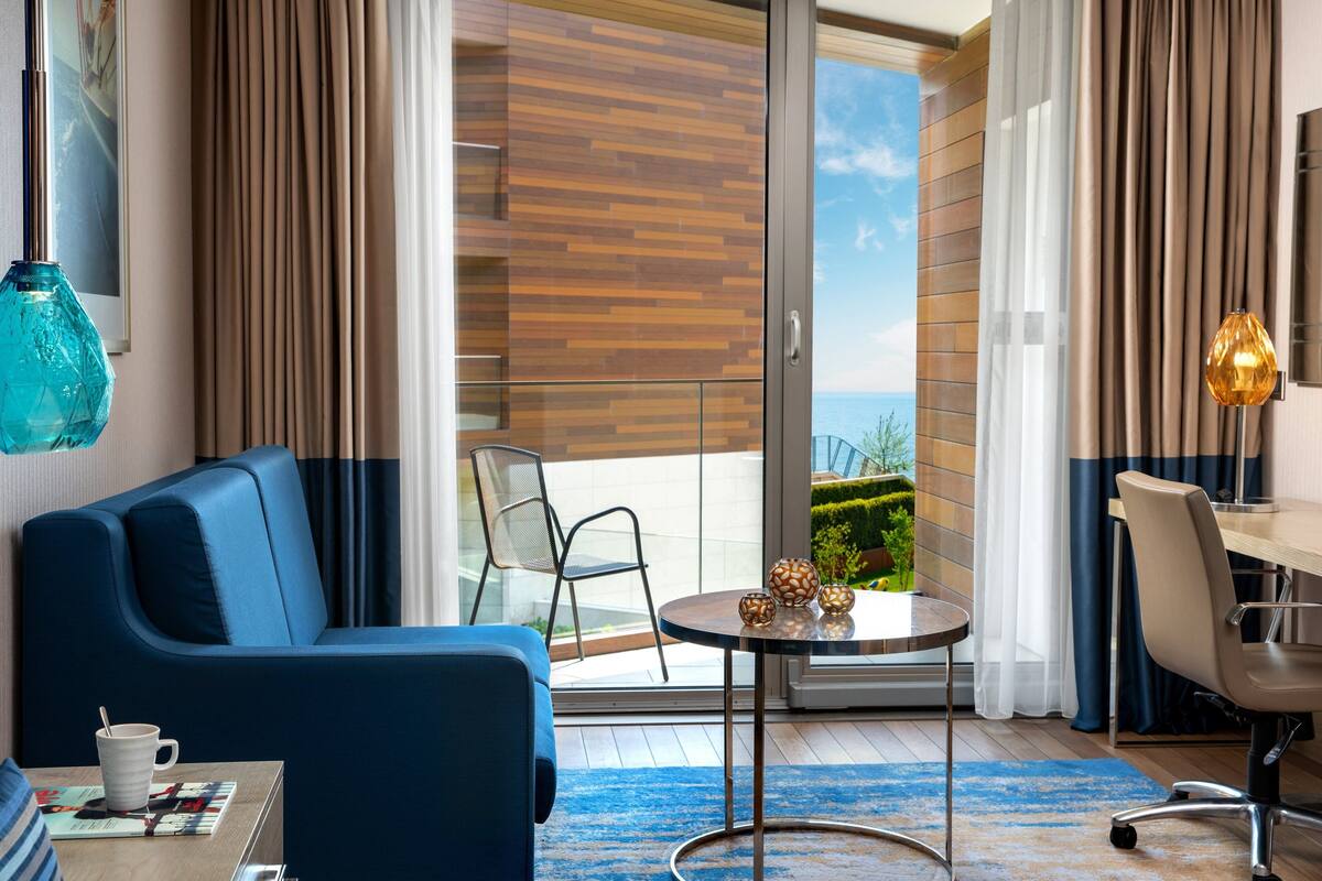 A modern hotel room with a blue sofa, a small round table with decorative items, a chair, and a view of the outdoors through a glass door.