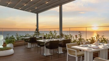 An outdoor dining area with tables and chairs, overlooking the sea at sunset.