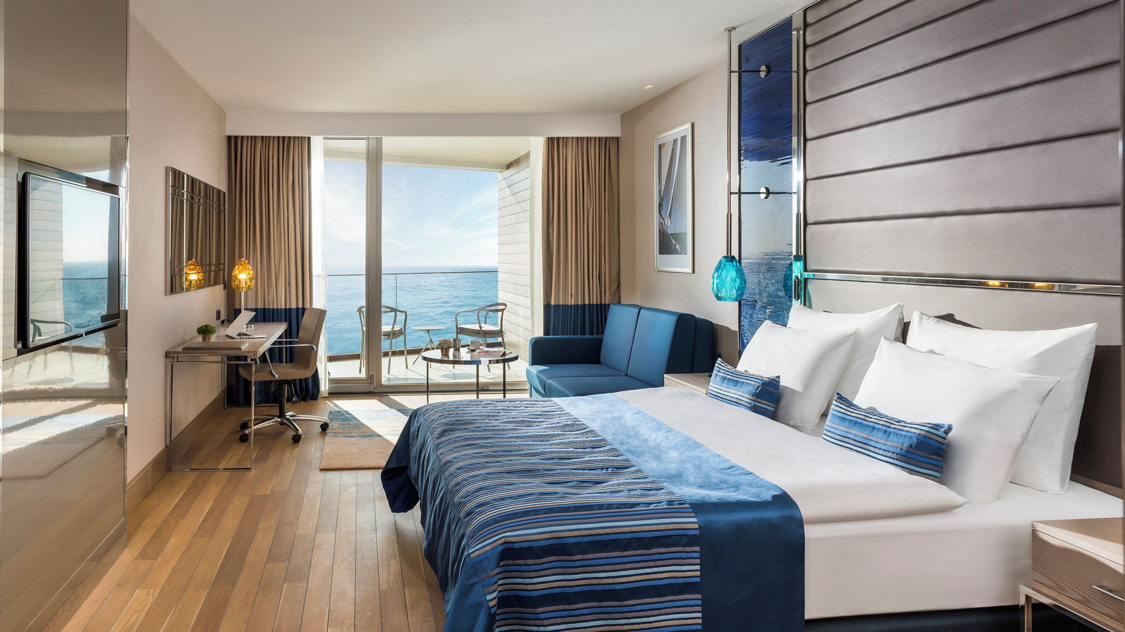 A modern hotel room with a large bed, a desk, a chair, and a sofa. There is a balcony with a view of the ocean.