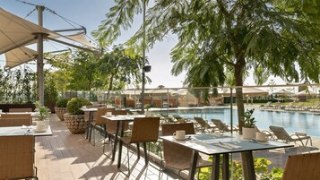 Un área de comedor al aire libre con mesas y sillas, una piscina y sombrillas.
