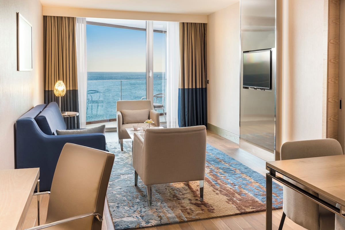 A hotel room with a large window, a sofa, a dining table, and a TV.