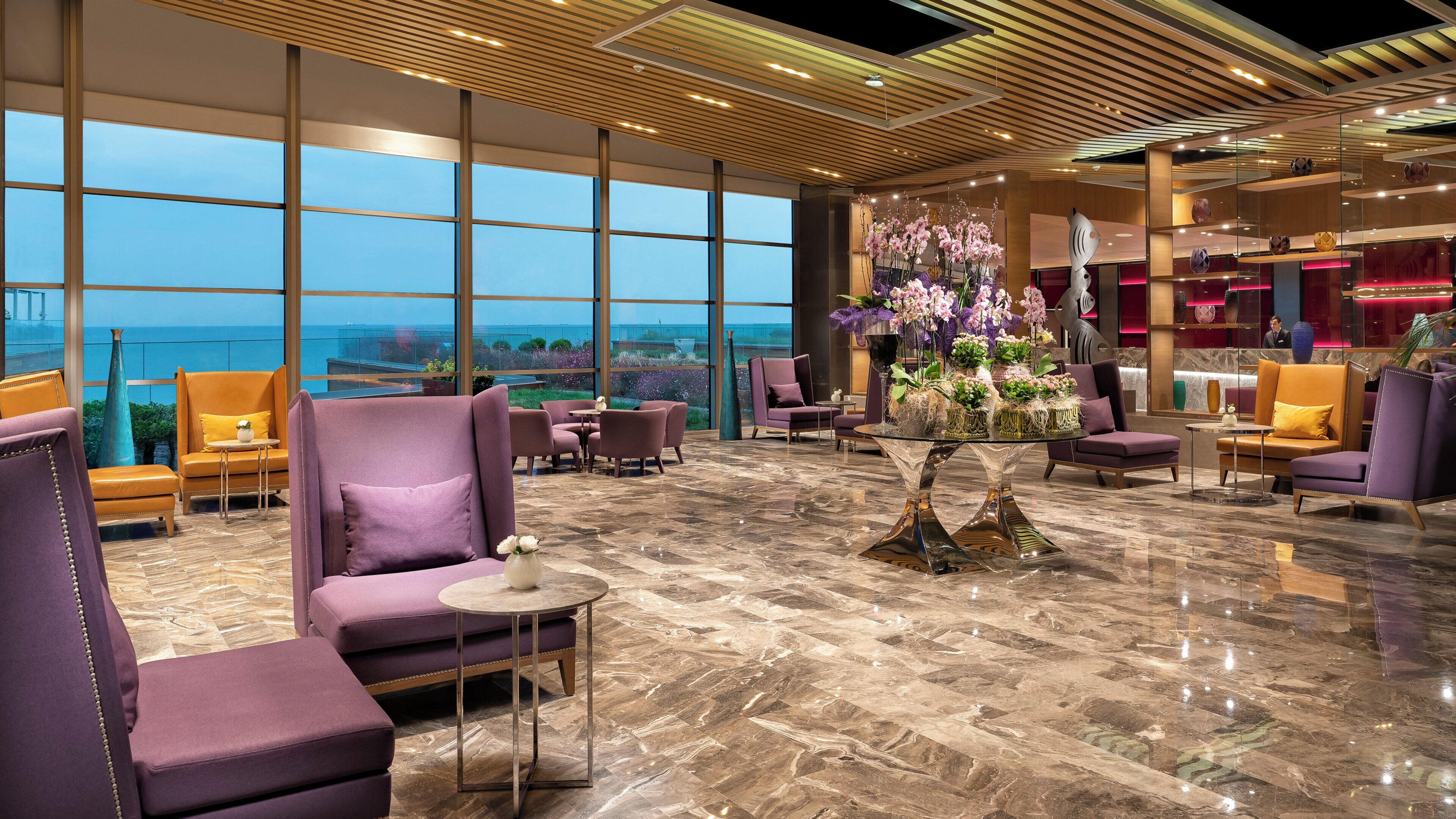 A spacious lobby with a large window offering a view of the sea, plush seating, and a central floral arrangement.