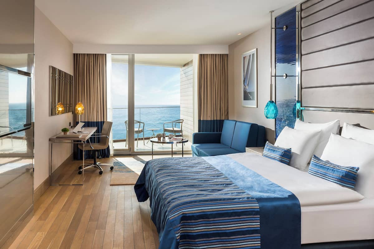 A modern hotel room with a large bed, a desk, a chair, and a sofa. There is a balcony with a view of the ocean.