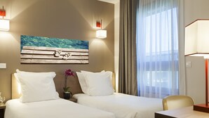 Une chambre d’hôtel avec deux lits, un tableau représentant un banc en bois sur lequel reposent des lunettes de soleil, une orchidée en pot et une vue sur un balcon à travers des rideaux transparents.