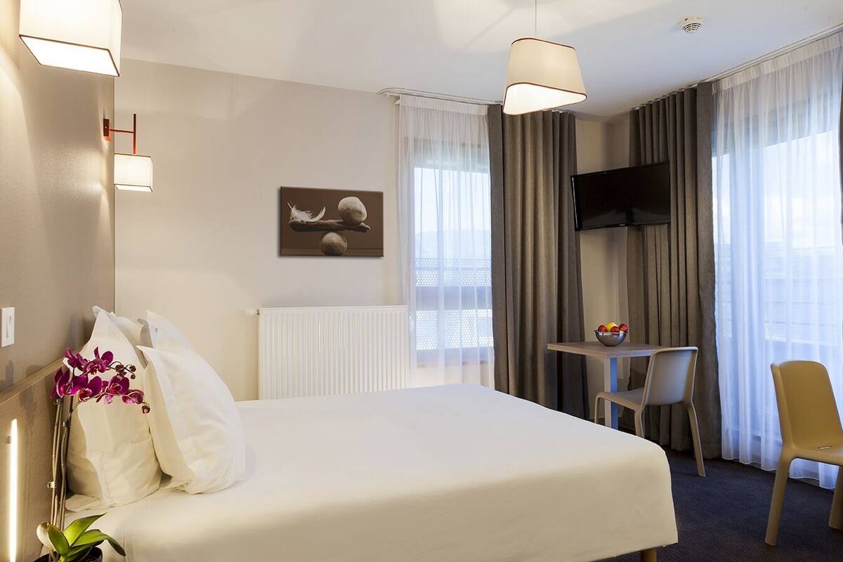Habitación de hotel con cama, escritorio, silla, televisor y ventana con cortinas.