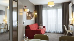 Une chambre d’hôtel avec un canapé rouge, une table à manger dressée avec des assiettes et des verres, un téléviseur fixé au mur et une grande fenêtre avec des rideaux.