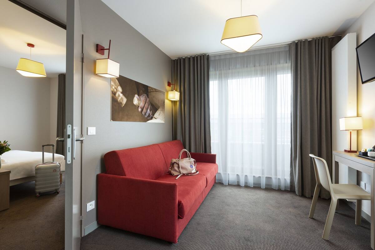 Habitación de hotel con sofá rojo, escritorio con silla, cama, maleta y un cuadro en la pared.