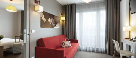 A hotel room with a red sofa, a desk with a chair, a bed, a suitcase, and a painting on the wall.