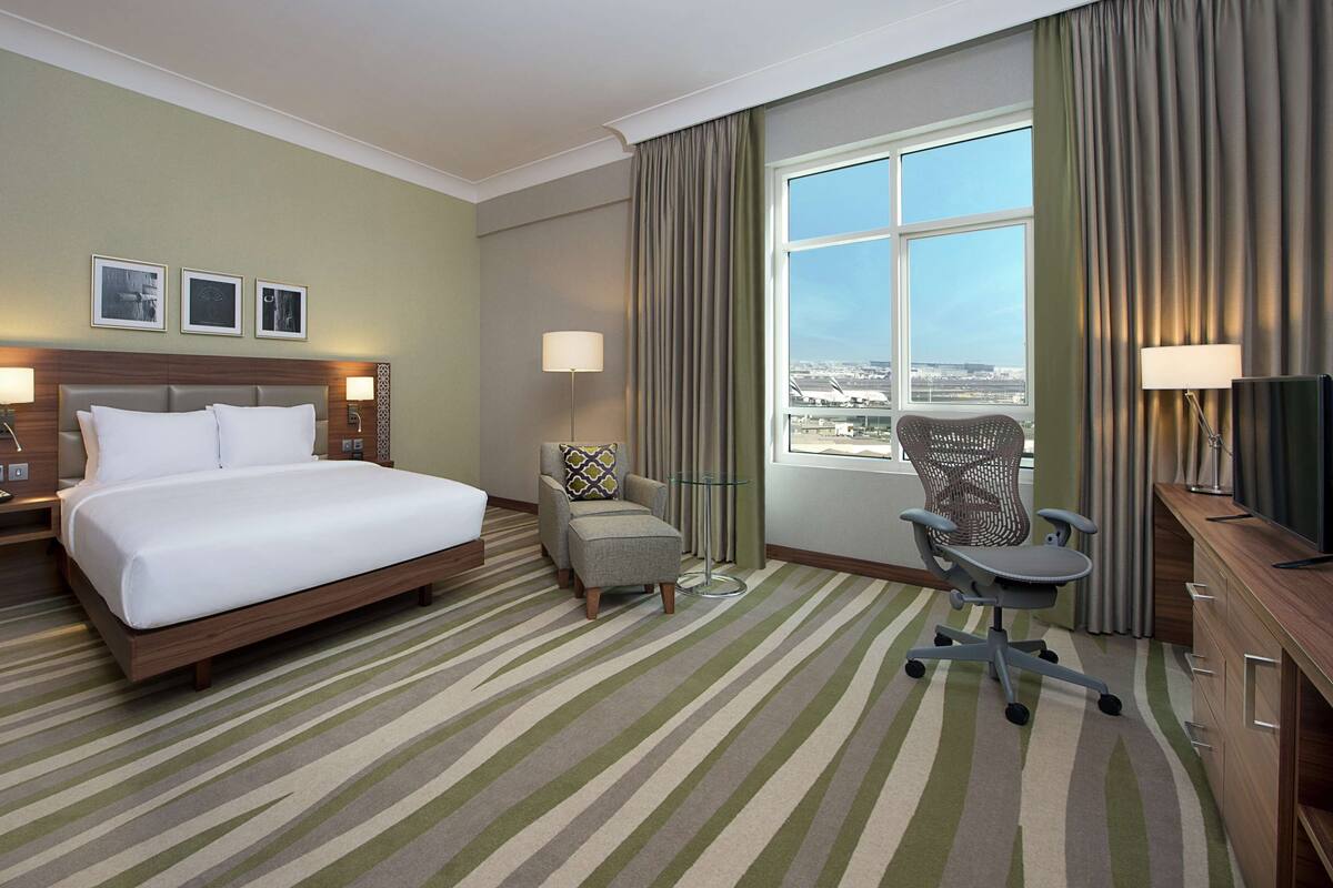 A hotel room with a large bed, a desk with a chair, a window with a view of the airport, and a striped carpet.