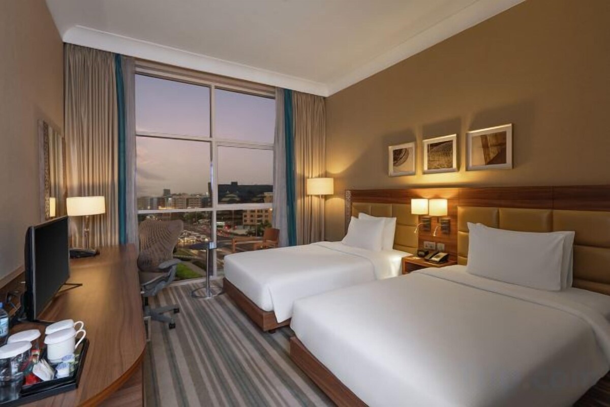 Une chambre d’hôtel avec deux lits, un bureau avec un ordinateur, une télévision et une fenêtre avec des rideaux.