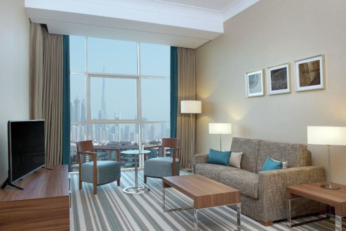 Quarto de hotel com janela ampla, sofá, televisão e área de refeições.