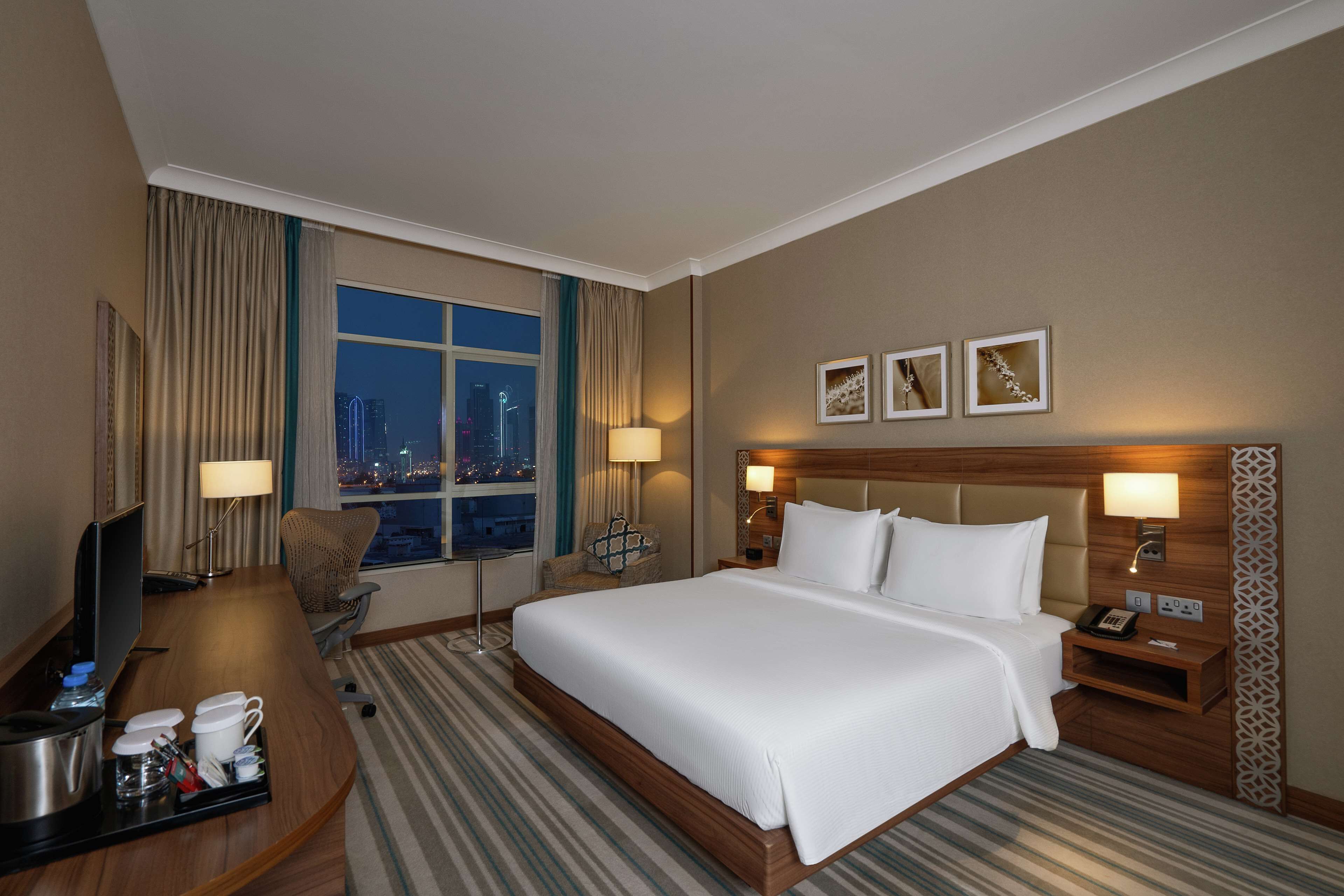 Habitación de hotel con una cama grande, un escritorio con televisor, una silla, un ventanal con vista a la ciudad y tres lámparas fijadas a la pared.