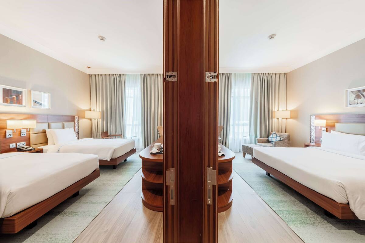 A hotel room with two beds, a wooden wardrobe, a round coffee table, and a large window with curtains.