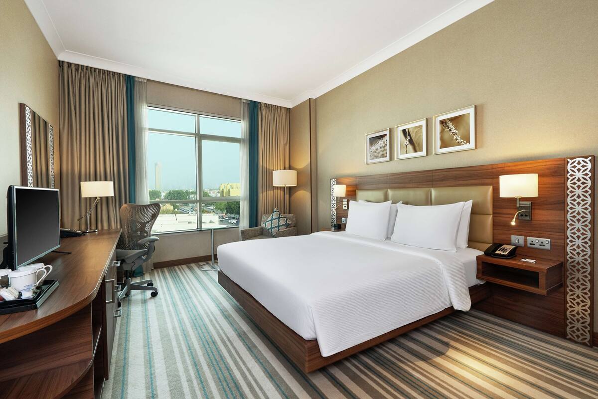A hotel room with a large bed, a desk with a computer, a chair, and a TV. There are two lamps on the bedside table and a window with curtains.