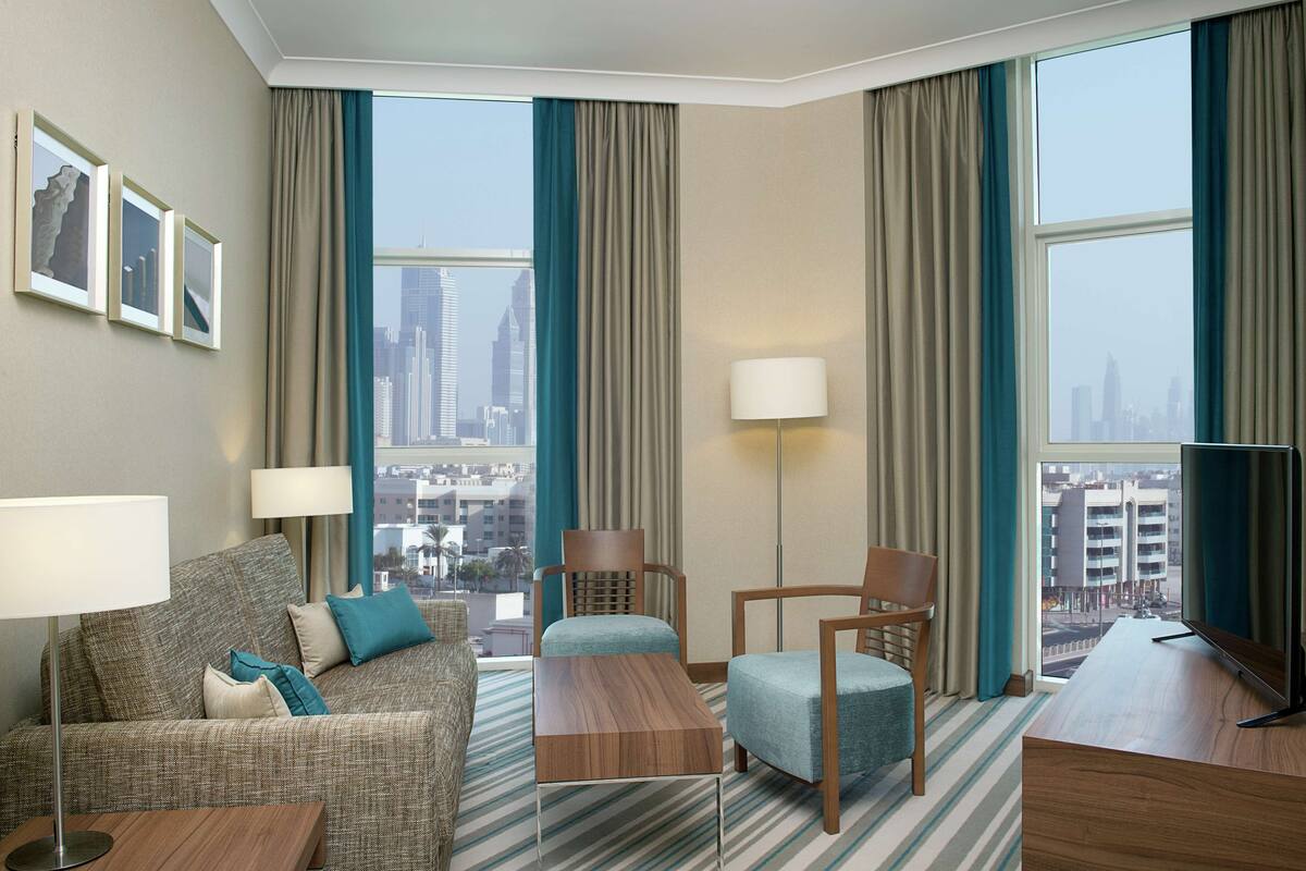 A hotel room with a sofa, two chairs, a coffee table, a TV, and large windows with a city view.