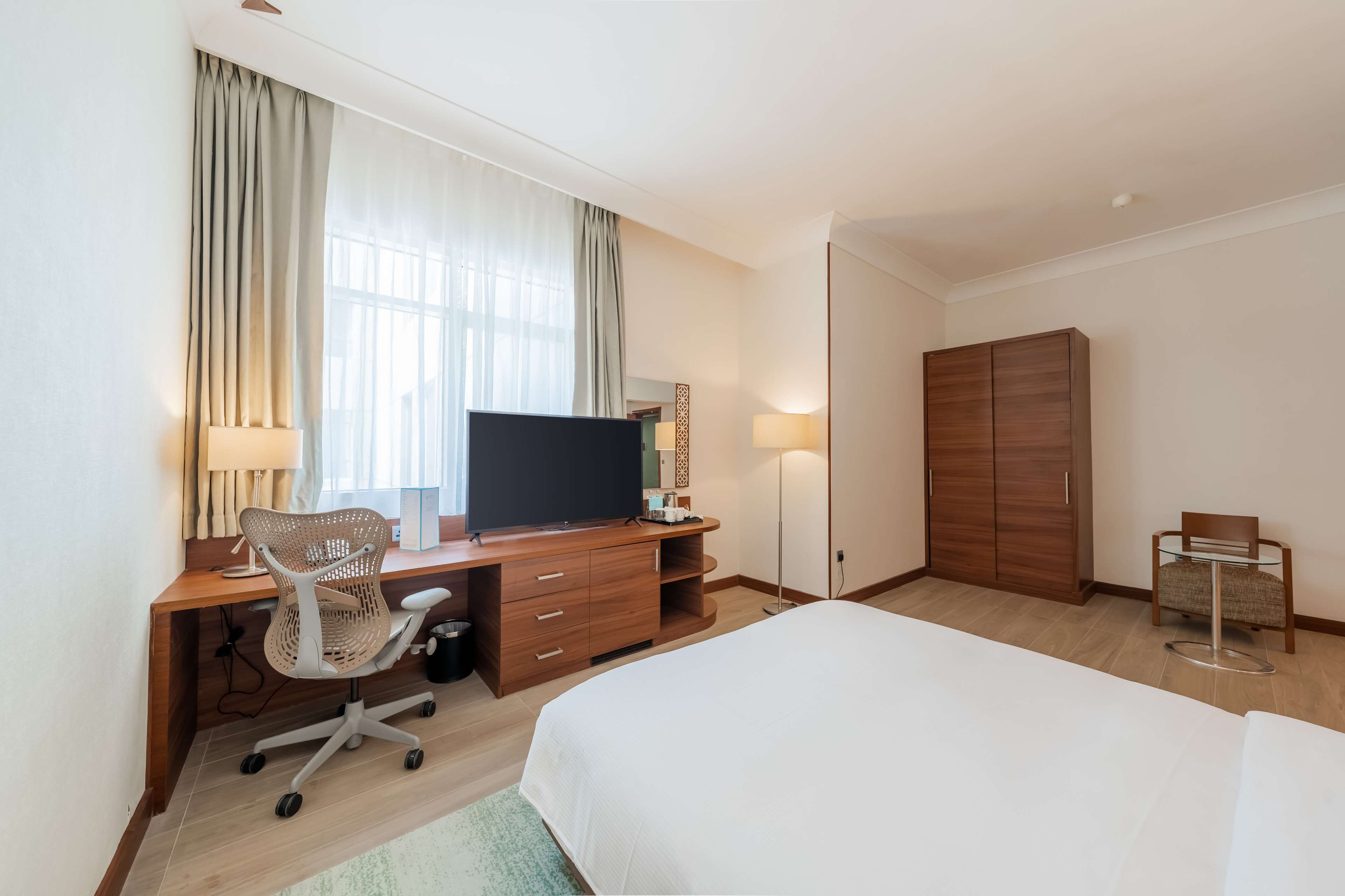 Una camera d'albergo con un letto, una scrivania con computer e sedia, una TV, un armadio e un tavolino con una poltrona.