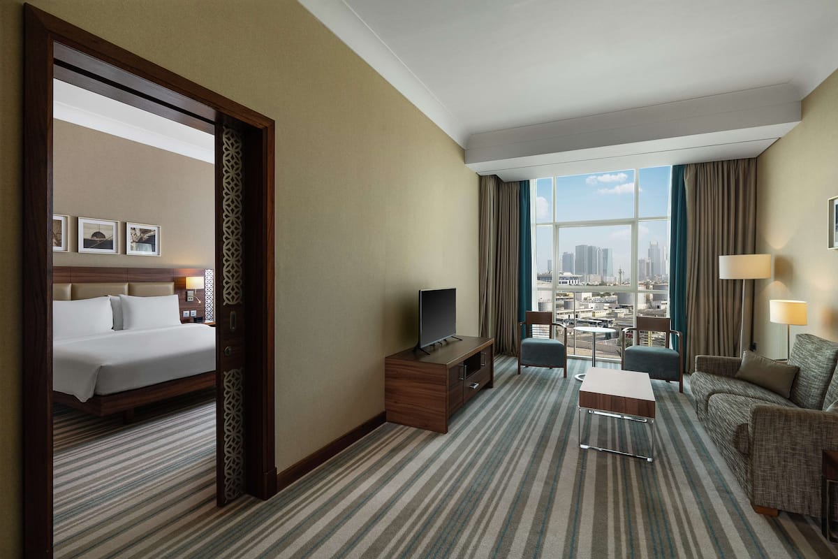 A hotel room with a bed, a TV on a wooden stand, a seating area with a sofa and chairs, and a large window offering a city view.