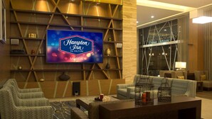 Un vestíbulo moderno con una gran pantalla de televisión mostrando 'Hampton Inn by Hilton Merida', asientos cómodos y una pared decorativa con estantes.