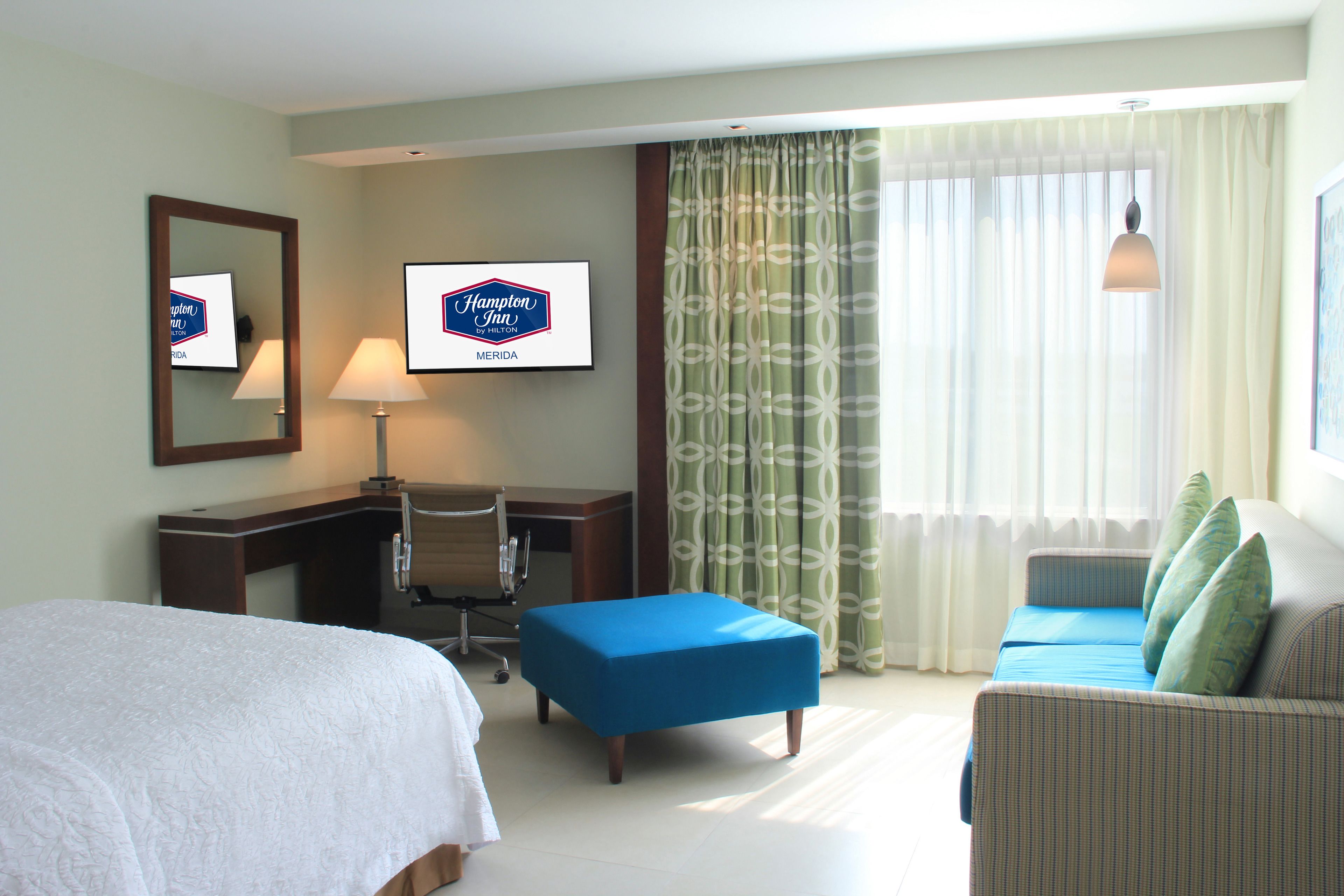 Ein Hotelzimmer mit Bett, Schreibtisch, blauem Hocker, einer Couch und einem großen Fenster mit Vorhängen.
