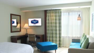 A hotel room with a bed, a desk, a blue ottoman, a sofa, and a large window with curtains.
