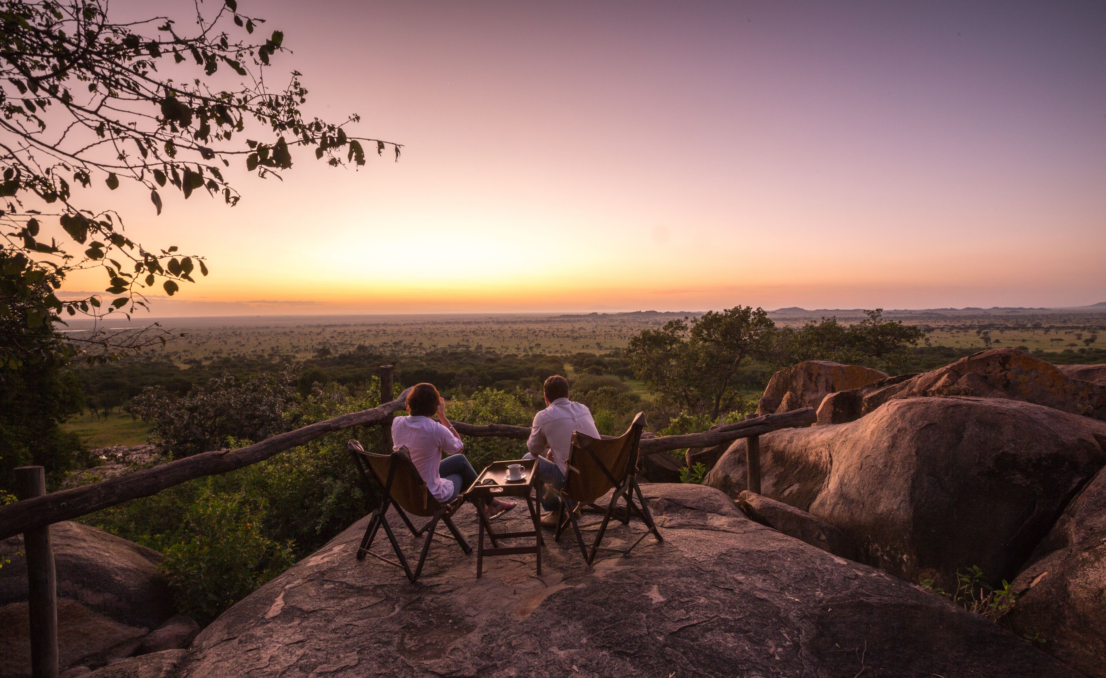 Two people sitting on chairs at a table with a view of a landscape during sunset.