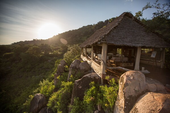 A rustic thatched-roof hut with a wooden deck, surrounded by lush greenery and large rocks. - Elewana Serengeti Pioneer Camp (Bariadi)