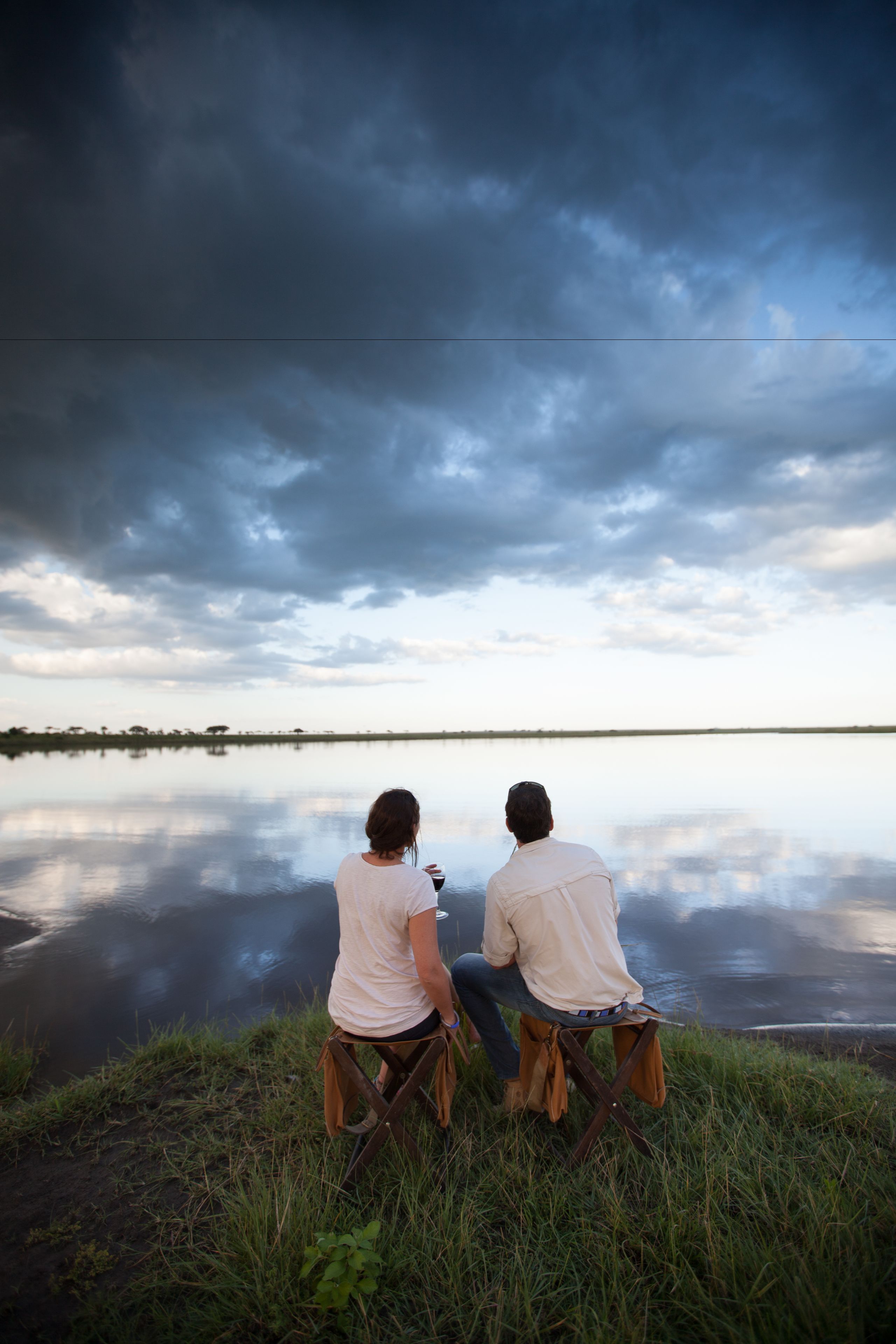 Two people sitting on chairs by a calm body of water with a cloudy sky above.