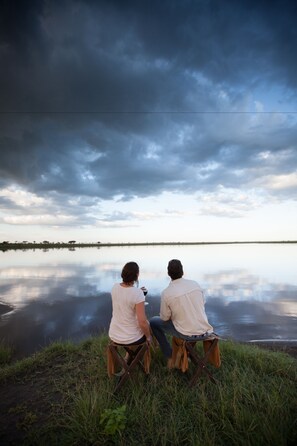 Two people sitting on chairs by a calm body of water with a cloudy sky above. - Elewana Serengeti Pioneer Camp (Bariadi)