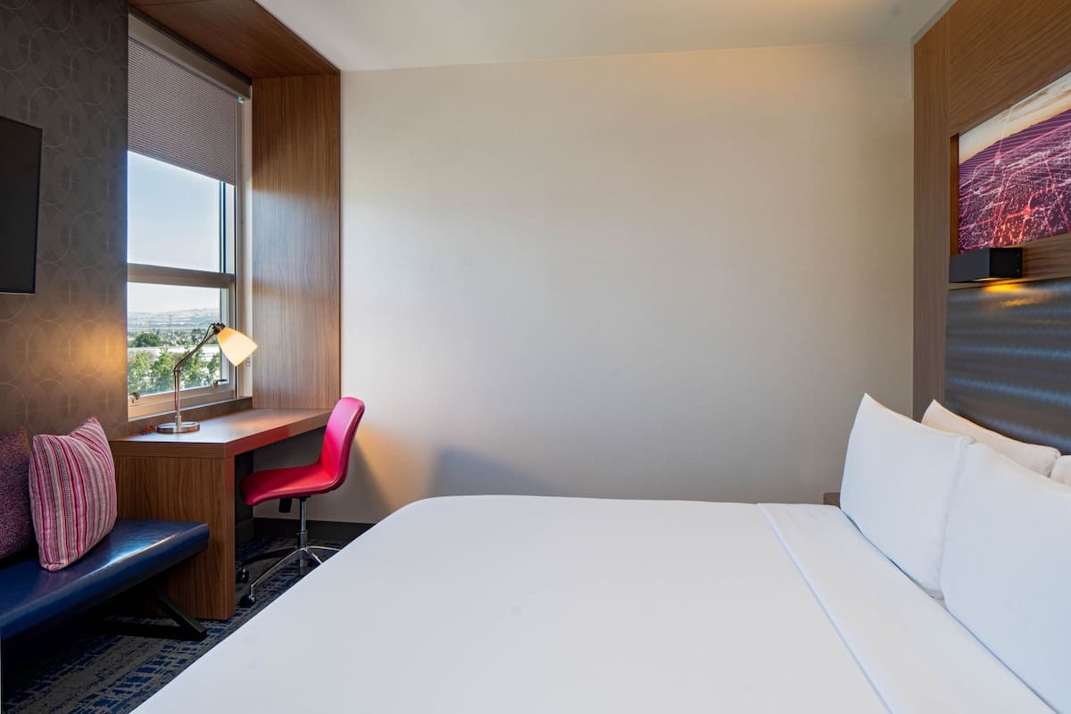 A hotel room with a bed, a desk with a chair, a window with blinds, a lamp, and a cityscape view.