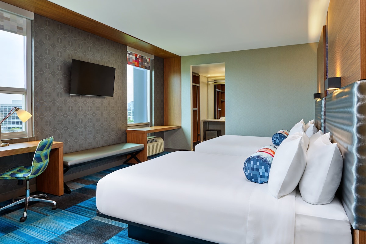 Ein Hotelzimmer mit einem großen Bett, einem Schreibtisch mit Stuhl, einem an der Wand befestigten Fernseher und Blick auf die Stadt.