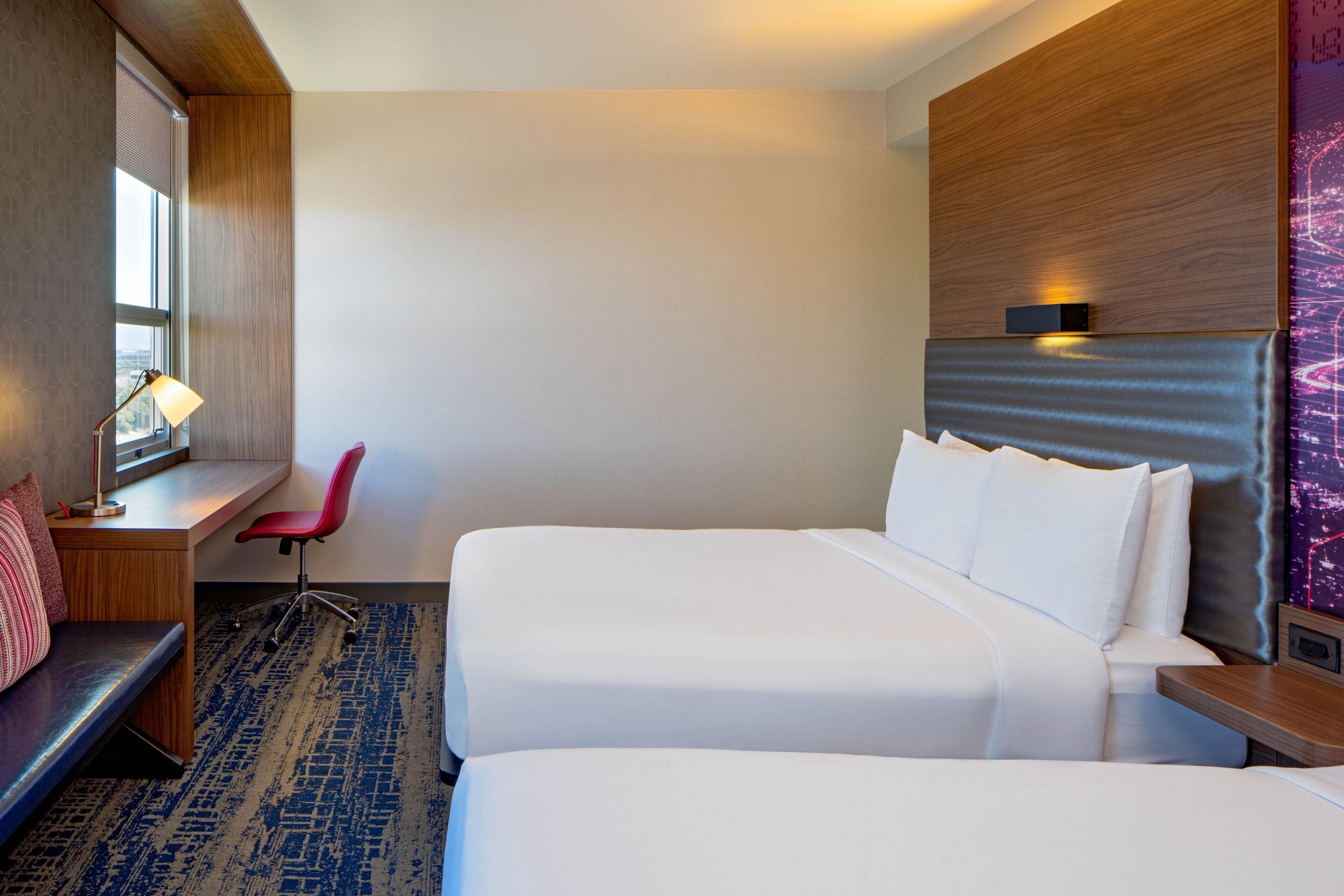 Quarto de hotel com duas camas, uma escrivaninha, uma cadeira e uma janela.