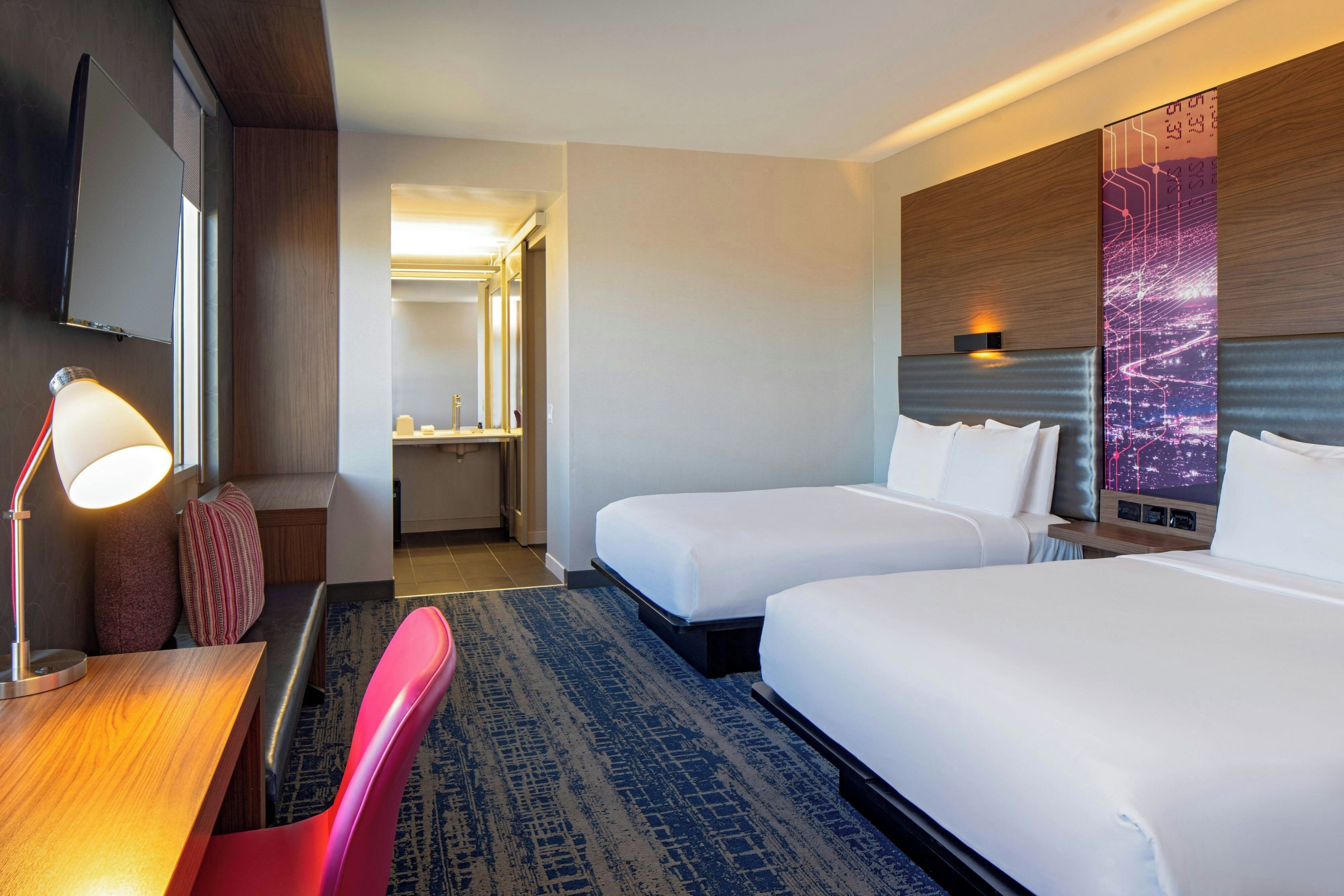 A hotel room with two beds, a desk with a lamp, a chair, a large mirror, and a wall-mounted TV.