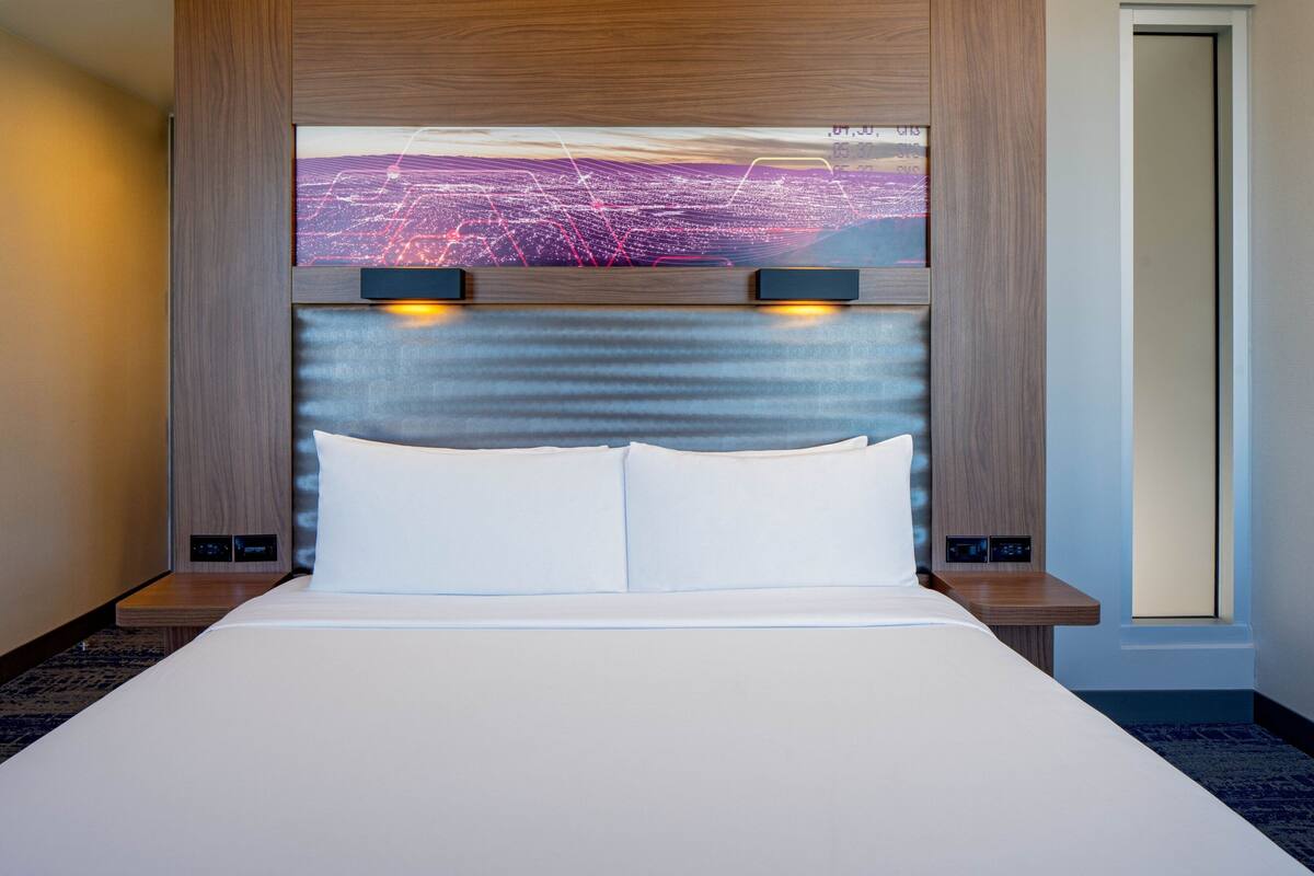 A hotel room with a large bed, a headboard with a cityscape artwork, and two bedside tables with lamps.