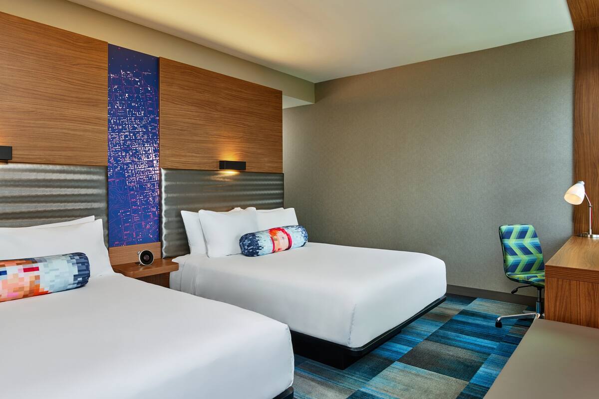 A hotel room with two beds, a cityscape wall art, a patterned rug, and a desk with a chair.