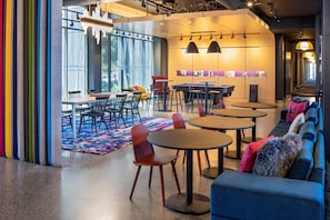 Relax in a stylish lounge area with a sofa, round tables, chairs, and a long bench, surrounded by artwork and a view of lush greenery.