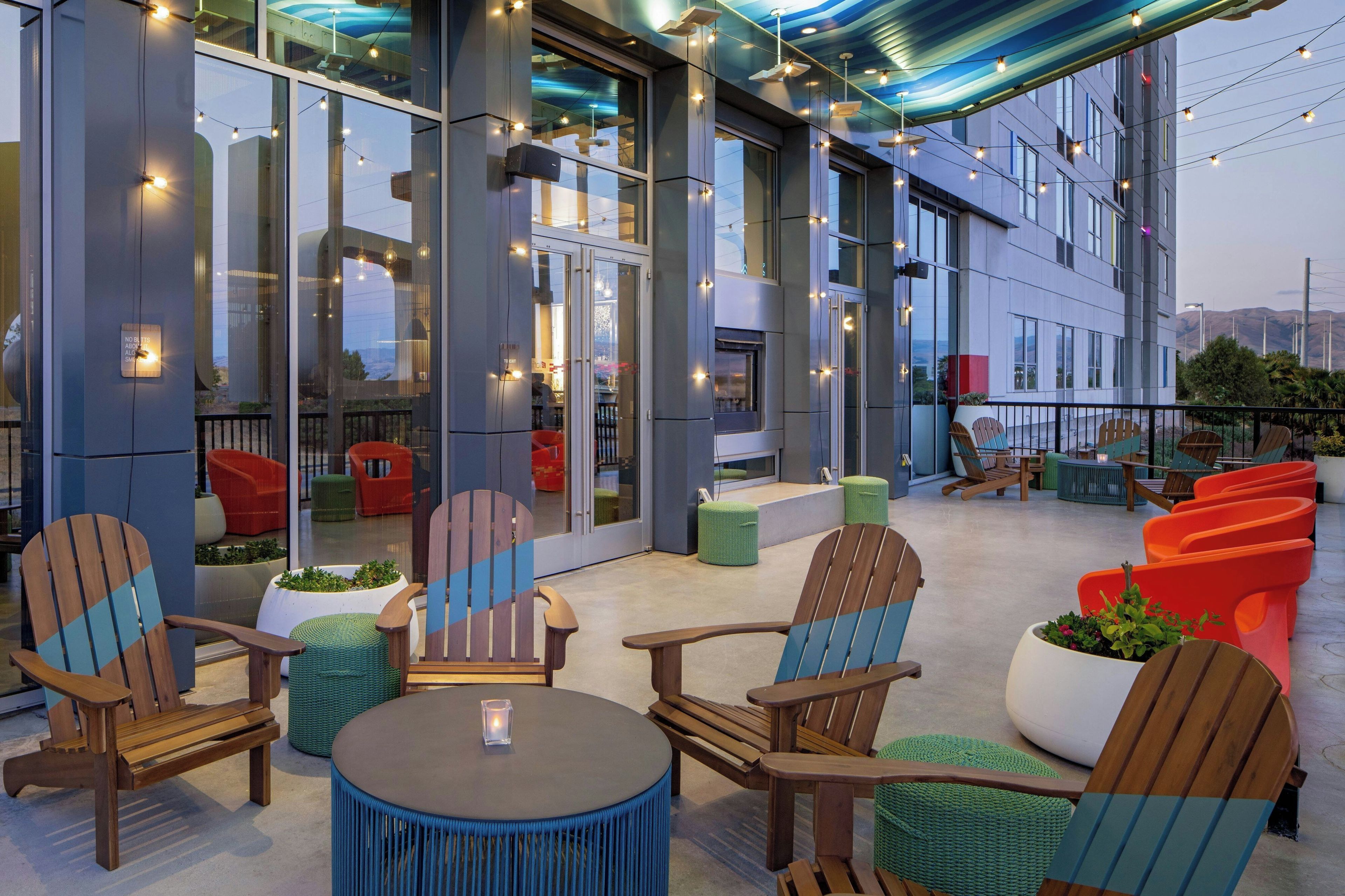 An outdoor seating area with wooden chairs and tables, a glass wall, and string lights.