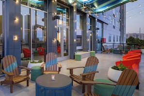 An outdoor seating area with wooden chairs and tables, a glass wall, and string lights. - Aloft by Marriott Santa Clara (San Jose)