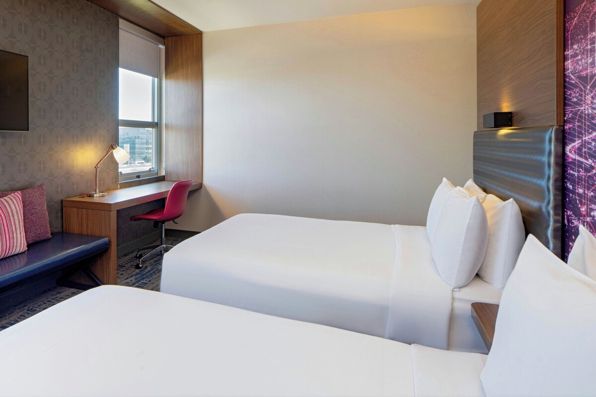 A hotel room with two beds, a desk, a chair, and a window.