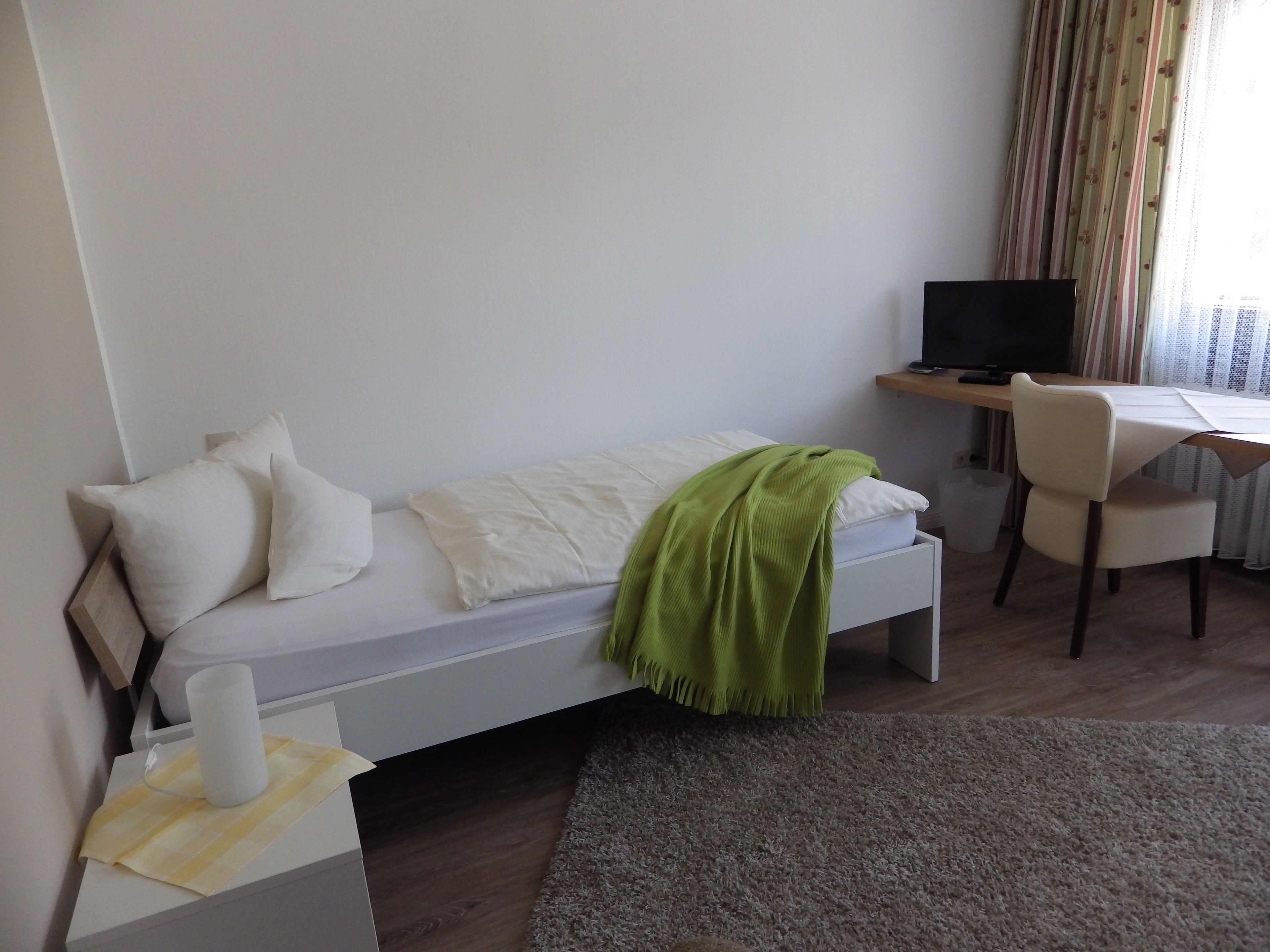 A small, neatly arranged bedroom with a single bed, a desk, a chair, and a television.