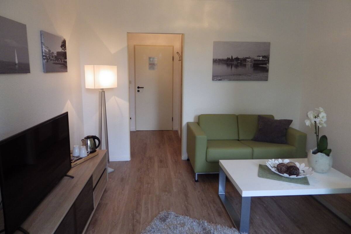 A living room with a flat-screen TV, a green sofa, a coffee table with a flower vase, and a door leading to another room.