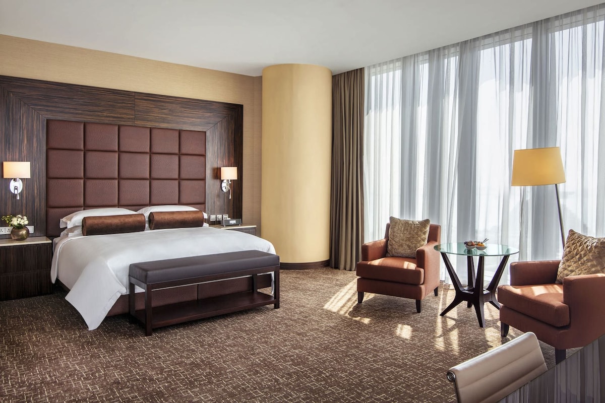 A hotel room with a large bed, a seating area with two armchairs and a small table, and a large window with curtains.