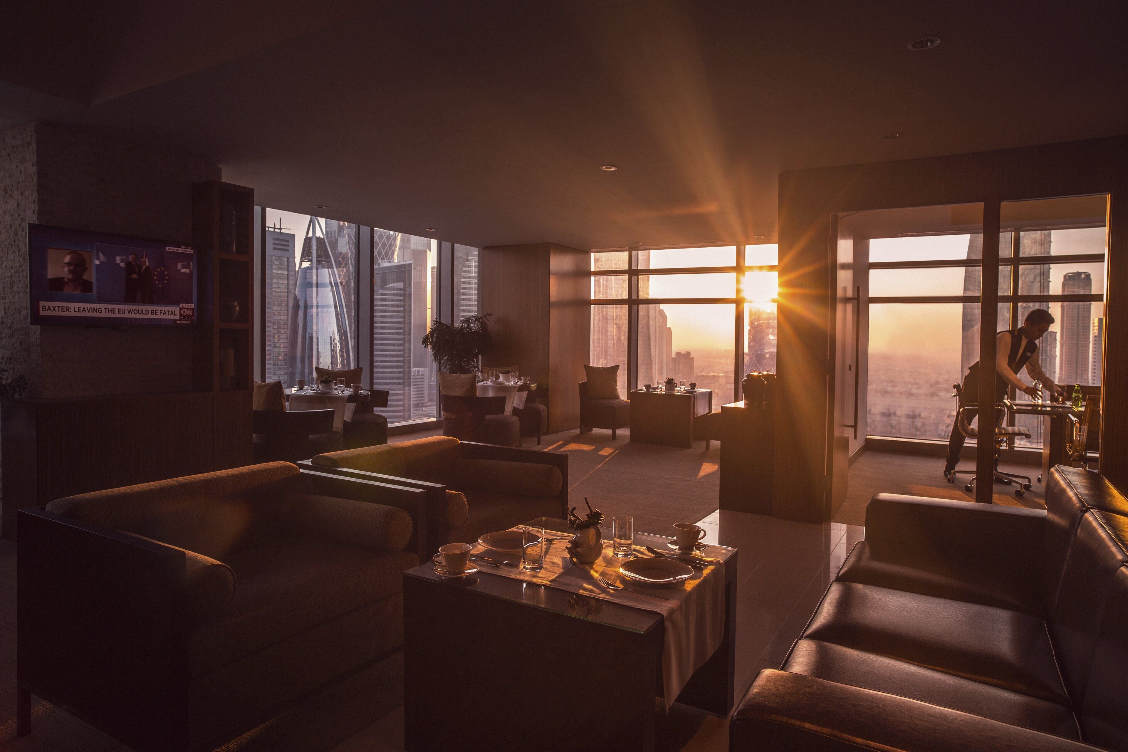 Habitación de hotel moderna con un amplio ventanal que ofrece vistas a la ciudad, un televisor de pantalla plana en la pared y una zona de comedor con una mesa para dos.