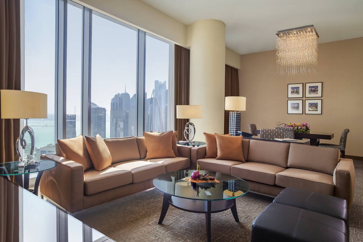 Habitación de hotel moderna con un ventanal grande que ofrece vista a la ciudad, una mesa de centro de vidrio, un sofá y una mesa de comedor con lámparas.