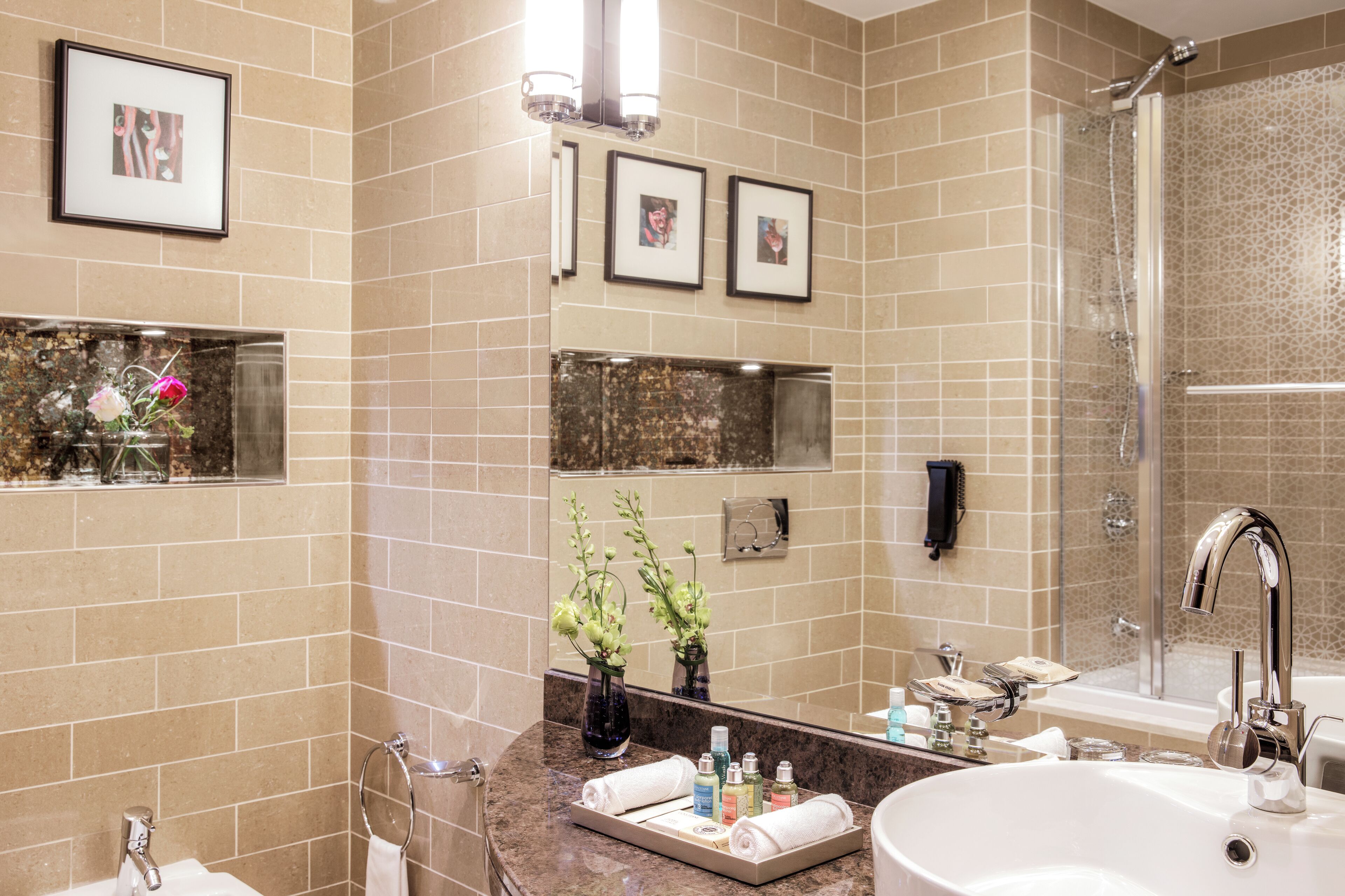 Un baño moderno con baldosas color beige, un espejo grande, cuadros enmarcados, un lavamanos con artículos de aseo y una zona de ducha.