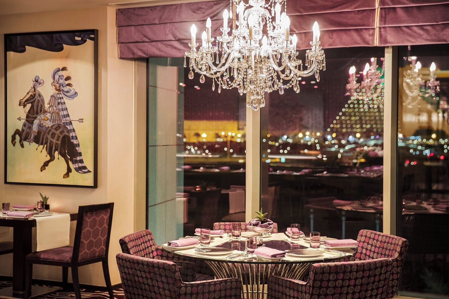 A dining area with a round table set for four, surrounded by patterned chairs. A large chandelier hangs above, and a cityscape is visible through the window.
